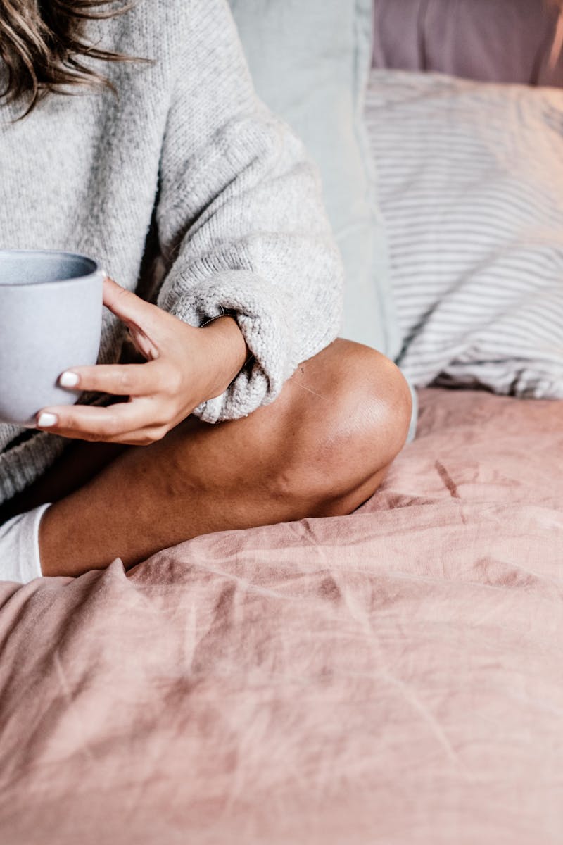 A woman enjoys a warm drink in bed, wearing a cozy sweater and socks. Intimate and relaxed atmosphere.